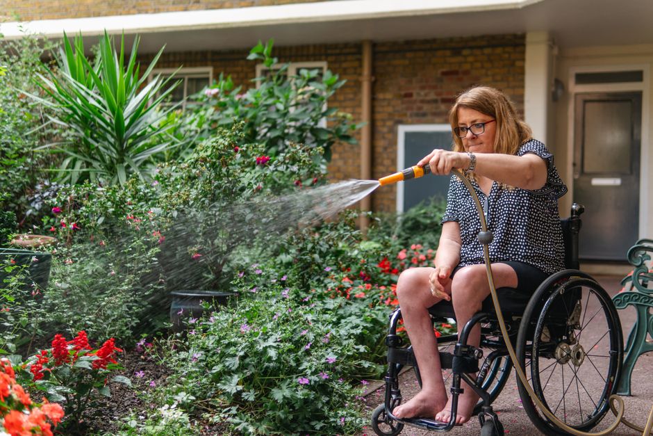 A woman in a wheelchair tending to a garden outside a brick residential building, watering vibrant flowering plants with a garden hose. The garden features a variety of lush green foliage, including t