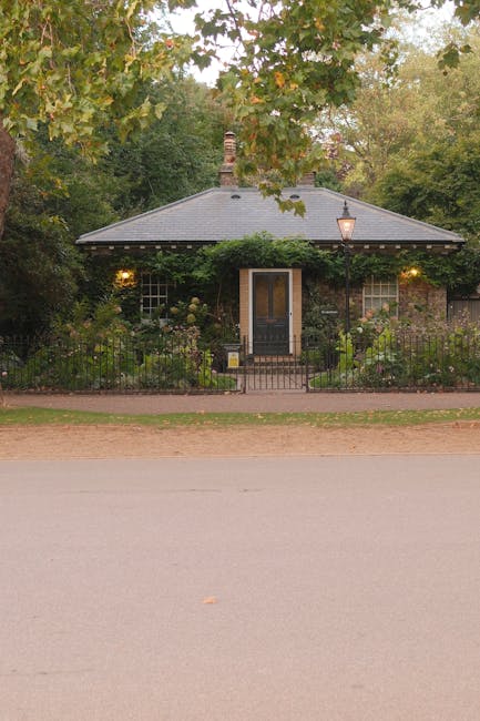 A small, single-story brick-fronted front garden of a cottage, enclosed by a low black metal fence with a central gate. The garden features a well-maintained flower border with various shrubs and bloo
