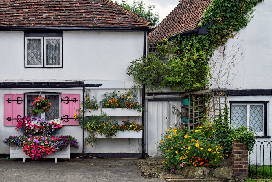 A small, single-story brick-fronted front garden of a cottage, enclosed by a low black metal fence with a central gate. The garden features a well-maintained flower border with various shrubs and bloo